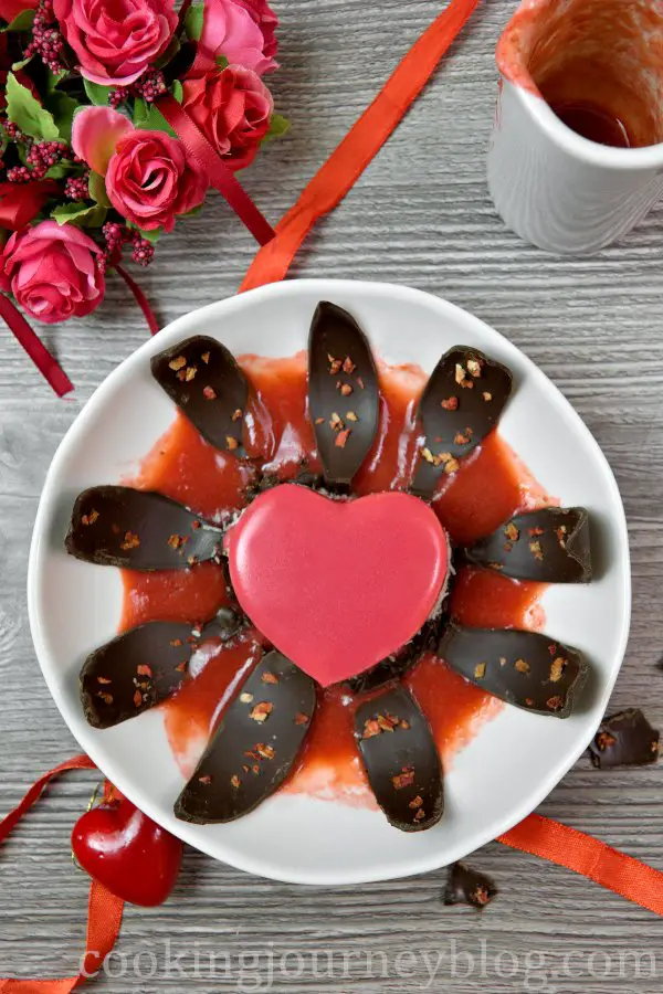 Plated Valentine's desserts, red mirror glaze cake on a plate with opwnwd chocolate leaves and strawberry desserts.