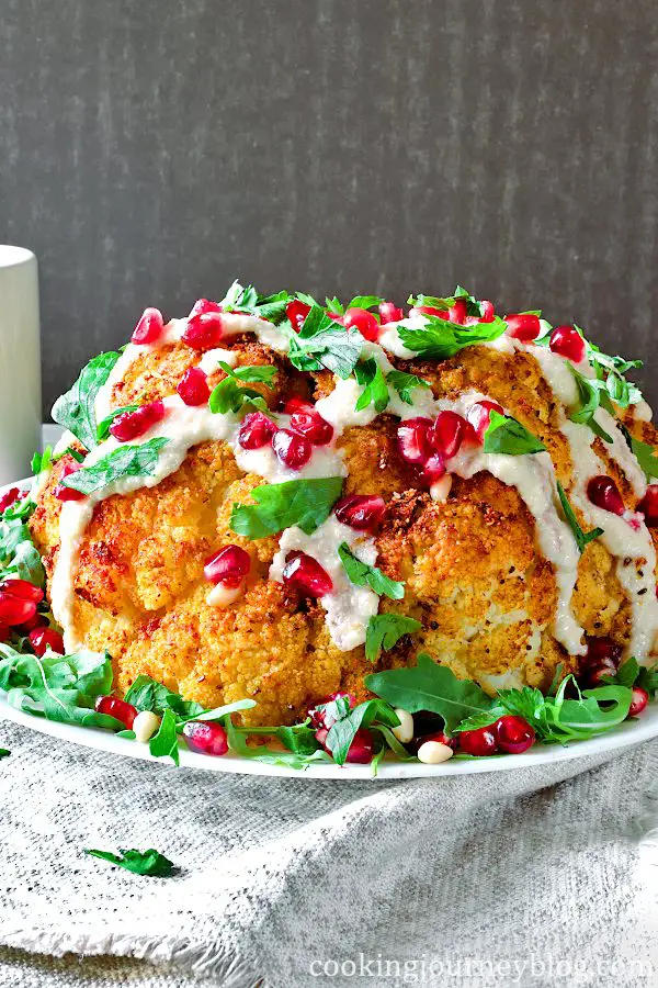 Whole roasted cauliflower with tahini sauce, pomegranate seeds and parsley. Served on a plate.