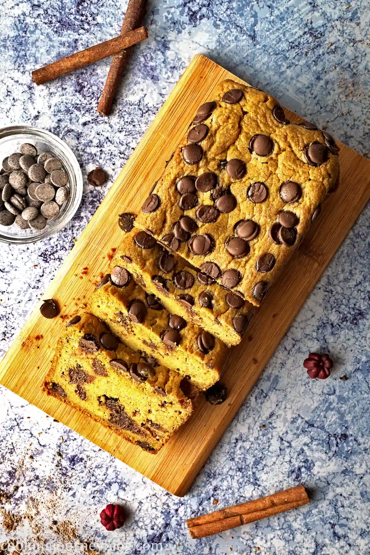 Half sliced pumpkin chocolate chip bread on a wooden board, served on a blue table with extra chocolate chips and cinnamon sticks.